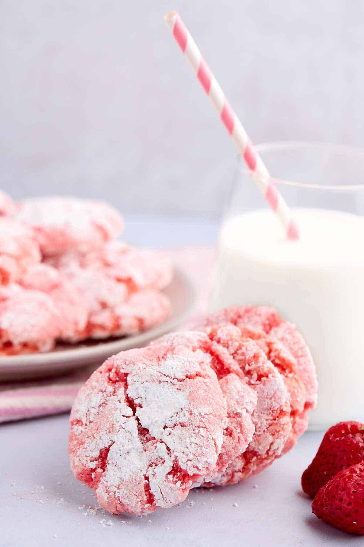 Strawberry crinkle cookies leaning against a cup of milk with a pink and white striped straw.