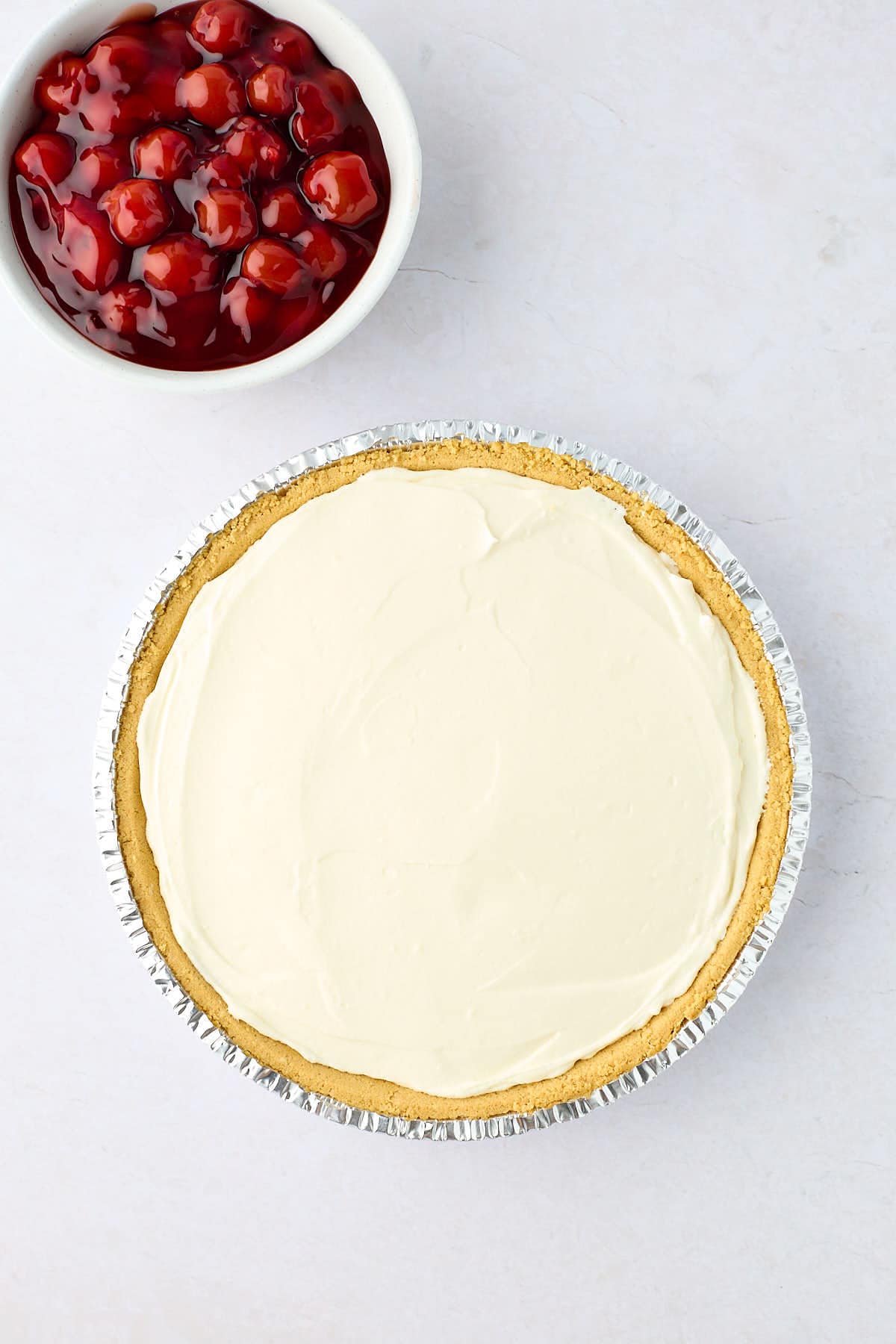 Cream cheese filling in a graham cracker pie crust with a bowl of cherry pie filling next to it.