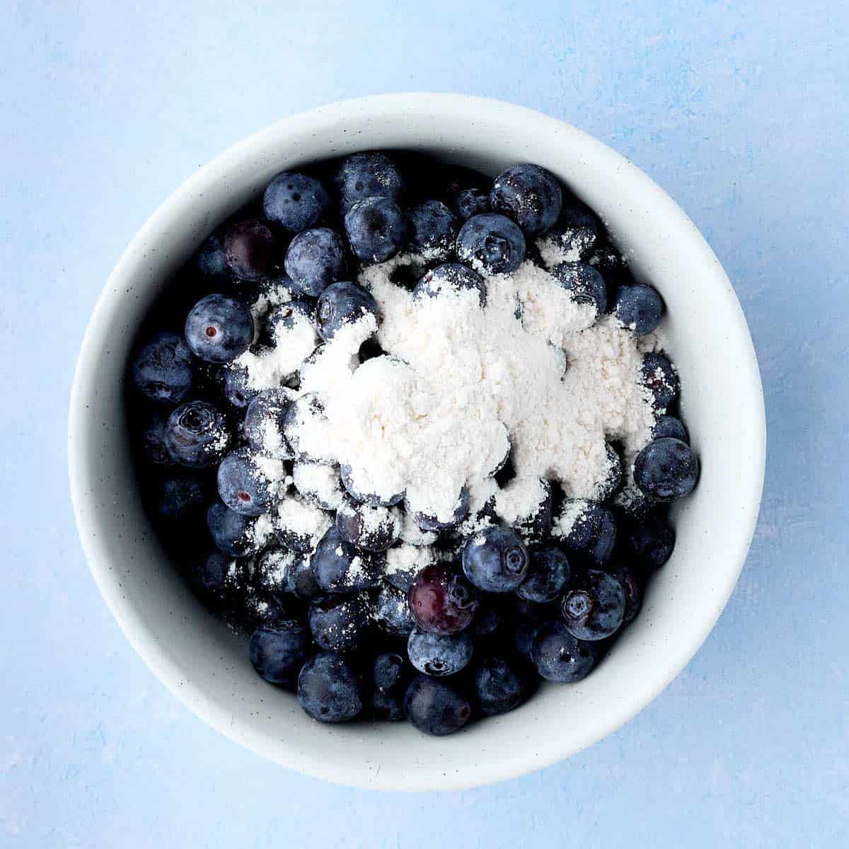 Bowl of fresh blueberries with a tablespoon of flour added, right before it is stirred to coat the blueberries.