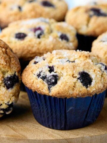 Closeup of a buttermilk blueberry muffin on a wooden board surrounded by other muffins.