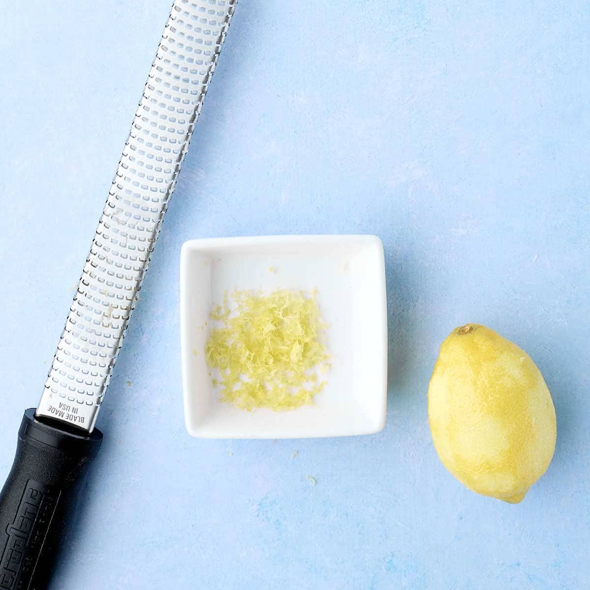 Zester, lemon zest in a bowl, and a lemon that has been zested lined up on the counter.